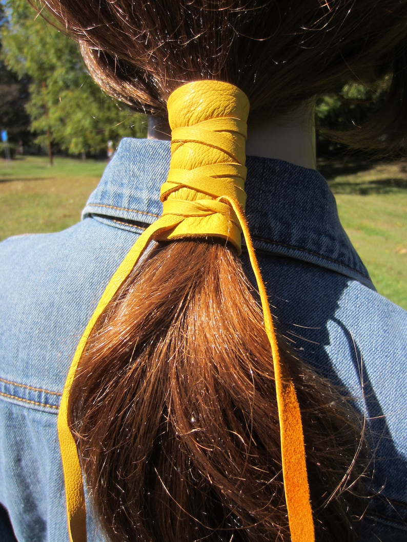 May include: A yellow leather ponytail holder with a braided design. The holder is tied around a brown ponytail.