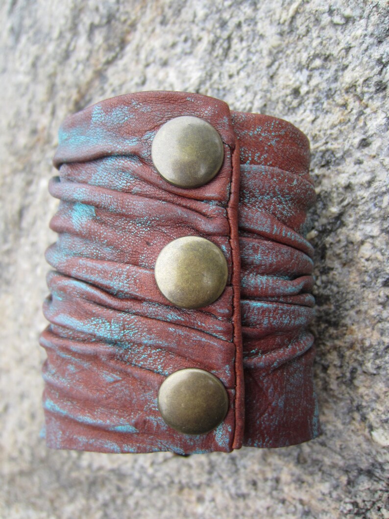 May include: A brown leather cuff bracelet with turquoise accents and three brass-colored metal snaps.