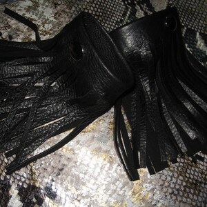 K&ouml;nnte beinhalten: Zwei schwarze Lederarmb&auml;nder mit Fransen. Jedes Armband hat einen silbernen Druckknopfverschluss.