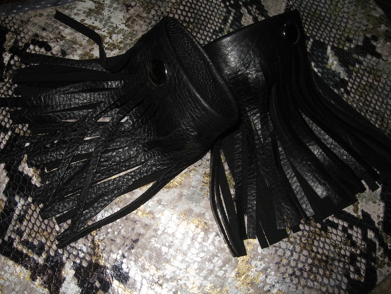 K&ouml;nnte beinhalten: Zwei schwarze Lederarmb&auml;nder mit Fransen. Jedes Armband hat einen silbernen Druckknopfverschluss.