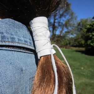 May include: White leather ponytail holder with a long white leather strap. The strap is tied around a ponytail and hangs down the back.