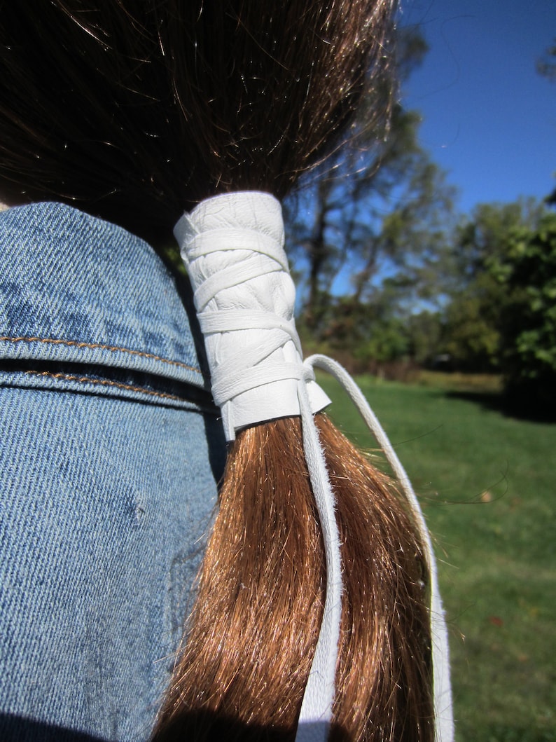 May include: White leather ponytail holder with a long white leather strap. The strap is tied around a ponytail and hangs down the back.