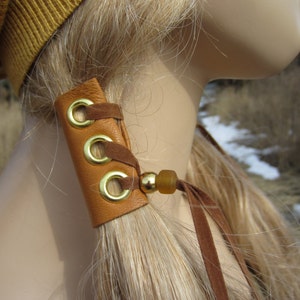 May include: A brown leather ponytail holder with three gold metal rings and a brown suede tie.