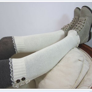 May include: A pair of legs are shown wearing gray tights and white knitted leg warmers with a lace trim and two brown buttons. The person is wearing gray lace-up boots.