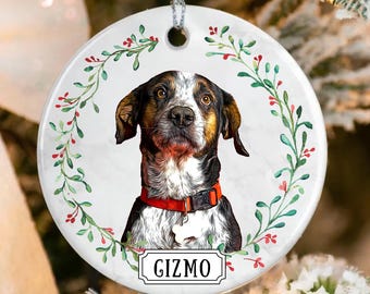 May include: A round, white ceramic Christmas ornament featuring a dog portrait with the name "GIZMO" below. The dog has brown, black, and white fur and wears a red collar. The ornament is decorated with a green and red floral wreath.