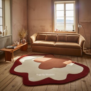 May include: A unique, abstract area rug with a red border, a white center, and a rust-colored accent. The rug is placed on a wooden floor in a room with a sofa and a window. The rug's design adds a modern touch to the room's decor.