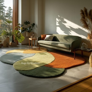 May include: A multi-coloured, abstract rug with an unusual, organic design. The rug features sections of green, yellow, orange, and grey. The rug is in a living room setting with a green sofa and plants. The text "Rugs By Bhadohi" is visible.