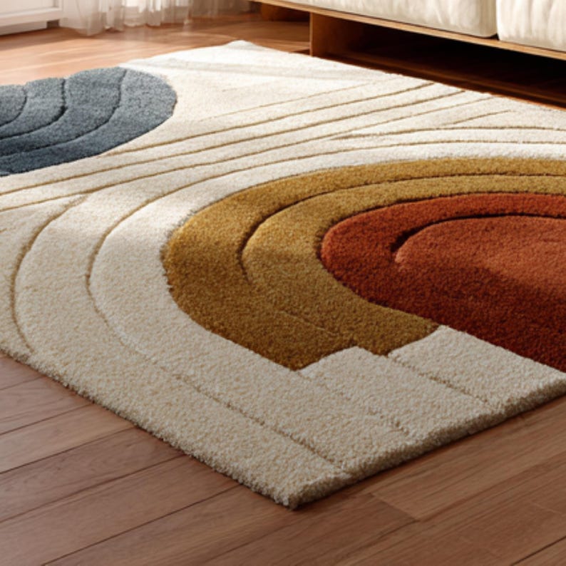 May include: A rectangular area rug with a modern design. The rug features a pattern of curved shapes in shades of cream, blue, gold, and rust. The rug is made of a soft, textured material and is placed on a wooden floor.