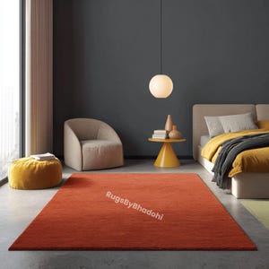 May include: A rectangular, rust-colored rug dominates the room's foreground. The rug is placed in a bedroom setting, with a neutral-toned chair, a yellow side table, and a bed with yellow bedding. The text "RugsByBhadohi" is visible on the rug.