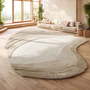 May include: A large, beige area rug with an unusual, organic shape. The rug has a layered design with varying shades of beige and tan, creating a textured, wave-like pattern. The rug is placed on a hardwood floor in a well-lit living space.