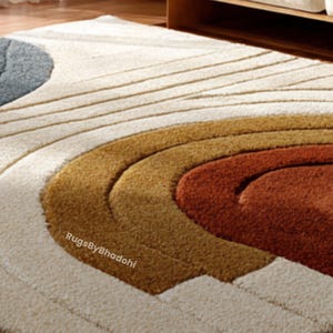 May include: A close-up of a beige and brown area rug with a circular design. The rug features concentric arcs in shades of beige, gold, and rust. The text "RugsByBhadohi" is visible in the lower left corner. The rug is on a wooden floor.
