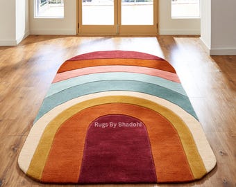 Op de afbeelding: Een regenboogvormig vloerkleed met gekleurde banden in rood, oranje, geel, groen en crème. Het kleed ligt op een houten vloer. De tekst "Rugs By Bhadohi" is zichtbaar op het kleed. Het ontwerp is kleurrijk.