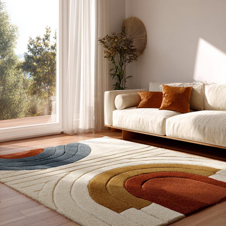 May include: A cream-colored area rug with a retro design featuring curved lines and semi-circles in shades of blue, brown, and rust. The rug is placed in a living room with a cream-colored sofa and rust-colored throw pillows. A large window and a decorative wall piece are also visible.