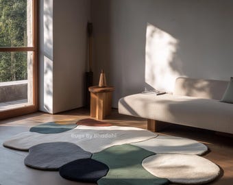 Puede incluir: Una alfombra de área abstracta multicolor con una forma orgánica única. La alfombra presenta varios tonos de verde, azul, gris, marrón y crema. La alfombra está colocada sobre un piso de madera en una habitación bien iluminada, con un sofá y una mesa auxiliar de madera en el fondo.
