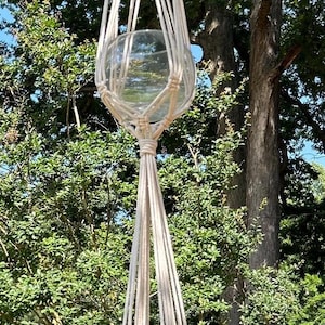 May include: A hanging macrame plant holder with two clear glass globes. The off-white macrame is suspended from a tree branch, with lush green foliage in the background. The globes are designed to hold plants.