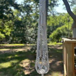 May include: A white macrame plant hanger with a wooden ring at the top. The plant holder is suspended from a tree branch, with a clear glass bowl inside. The background is a blurred view of green trees and grass.