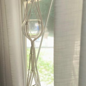 May include: A white macrame plant hanger with a clear glass orb and a small ceramic pot holding a green plant. The hanger is suspended in front of a window with white curtains.