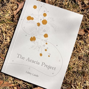 May include: A book titled "The Acacia Project" by Libby Lamb. The cover displays a botanical illustration of yellow flowers and leaves with dotted lines and small stars. The book rests on a bed of dry grass and leaves.