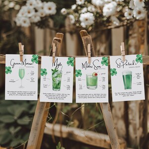 May include: Four recipe cards for St. Patrick's Day cocktails and beer are displayed on a wooden stand. The cards are held by clothespins and feature illustrations of drinks like a Green Mimosa and a Mojito, with ingredients listed.