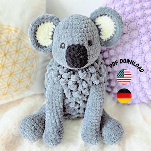 May include: A gray crocheted koala plush toy with a large black nose, round ears with white centers, and dark eyes. The text PDF DOWNLOAD is visible next to the koala, accompanied by a United States flag icon and a German flag icon.
