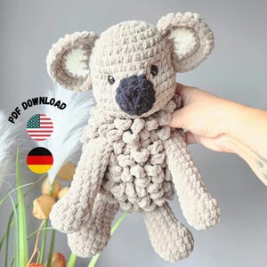 Könnte beinhalten: Ein hellbraunes, gehäkeltes Koala-Plüschtier mit einer dunkelgrauen Nase, großen, abgerundeten Ohren mit weißer Mitte und einem strukturierten Garnkörper. Das Bild enthält den Text PDF DOWNLOAD sowie amerikanische und deutsche Flaggensymbole.