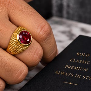 Puede incluir: Un anillo de tono dorado con una gran piedra roja redonda y facetada, engastada en una banda texturizada con superficie granulada. Junto al anillo hay un objeto rectangular negro con el texto dorado: BOLD CLASSIC PREMIUM ALWAYS IN STYLE.