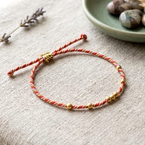May include: A red, pink, and gold braided bracelet with gold beads. The bracelet is adjustable and has a delicate, handcrafted appearance. The bracelet is a piece of jewelry.