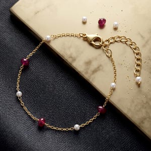 May include: A gold chain bracelet featuring alternating small white pearls and faceted deep red ruby beads. The bracelet includes a lobster claw clasp and an adjustable extender chain with a single pearl at the end, showcasing a delicate design.