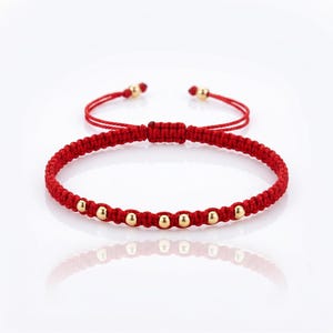 Puede incluir: Pulsera de macramé roja con cuentas doradas. La pulsera tiene un cierre de nudo corredizo para ajustar la talla. Las cuentas están espaciadas uniformemente a lo largo del cordón rojo.