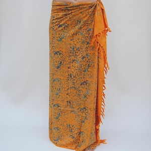 Könnte beinhalten: Ein orangefarbenes Sarong mit einem blau-orangen Blumenmuster. Das Sarong ist um die Taille gewickelt und an der Seite gebunden. Der Rand des Sarongs hat Fransen. Das Sarong ist aus leichtem Stoff gefertigt.