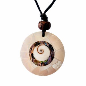 Könnte beinhalten: Ein runder Anhänger, bestehend aus einem Mosaik aus schillernden weißen Muschelstücken, die einen dunklen, schimmernden Abalone-Ring und ein kleines weißes Spiralmuschel-Zentrum umgeben, befestigt an einer schwarzen Kordel mit einer braunen Holzperle.