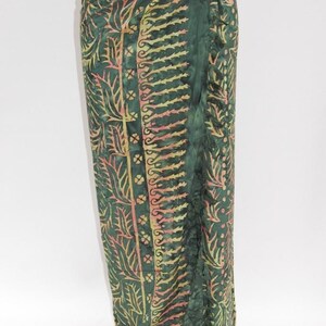 Può includere: Un sarong verde lungo con un motivo in stile batik. Il tessuto presenta un motivo ripetuto di foglie ed elementi floreali nelle tonalità del giallo e del rosa. Il sarong ha un bordo a balze ed è legato in vita.