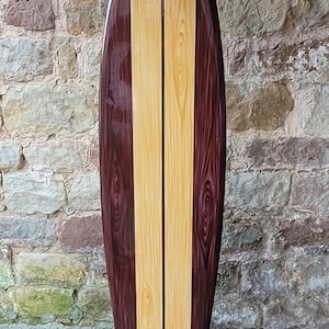 May include: A wooden surfboard with a glossy finish. The board features a vertical design with two light-colored wood panels flanked by darker wood panels. It is displayed on a black metal stand, against a stone wall.