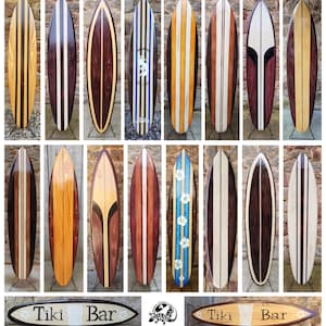 Può includere: Una collezione di tavole da surf in legno con vari design, tra cui strisce e colori uniti. Le tavole da surf sono esposte contro un muro di mattoni, mettendo in risalto la loro maestria artigianale e il loro fascino estetico.