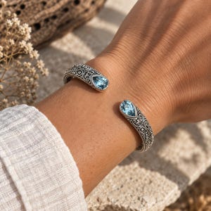 Puede incluir: Una pulsera de brazalete abierto hecha de plata oxidada con intrincados patrones de filigrana y dos piedras preciosas de color azul claro en forma de l&aacute;grima engastadas en cada extremo, creando un aspecto equilibrado y detallado.