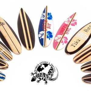 Könnte beinhalten: Eine Sammlung von elf Miniatur-Surfbrett-Ornamenten aus Holz, die fächerförmig angeordnet sind. Die Bretter zeigen verschiedene Designs, darunter Streifen in Naturholzoptik, blaue und rosa Hibiskusblütenmuster sowie die Schriftzüge Surfer Boy, Surfer Girl und Tiki Bar.