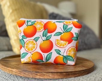 May include: A small, zippered pouch with a repeating pattern of oranges, orange slices, green leaves, and white blossoms on a light pink background. The pouch is sitting on a round, wooden tray.