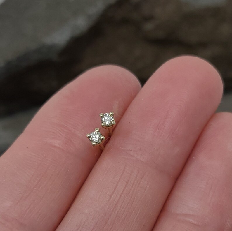 May include: A pair of small, gold-colored stud earrings, each featuring a single, clear, round-cut gemstone. The earrings are set in a four-prong setting, and are displayed against a blurred background.