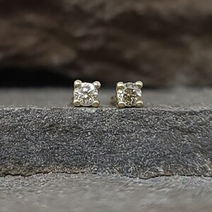 May include: A pair of small, square-shaped stud earrings. Each earring features a clear, round gemstone set in a gold-colored metal. The earrings are displayed on a dark gray, textured surface.