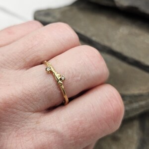 May include: A gold ring with a textured, branch-like band featuring two small, clear, round-cut stones set along the top. The metal has a matte, organic finish that mimics the appearance of a twig, with the stones secured in raised settings.