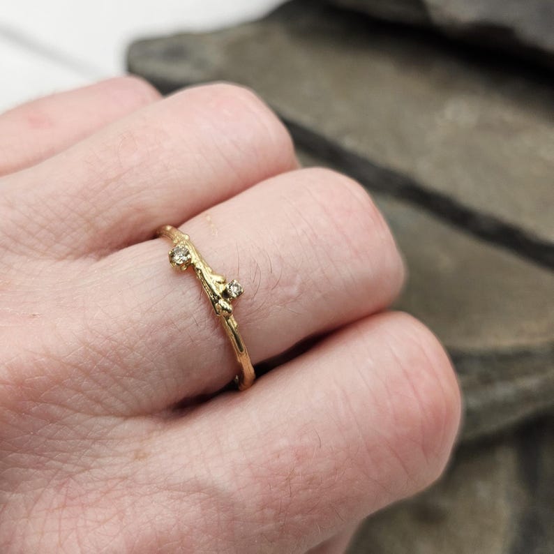 May include: A gold ring with a textured, branch-like band featuring two small, clear, round-cut stones set along the top. The metal has a matte, organic finish that mimics the appearance of a twig, with the stones secured in raised settings.