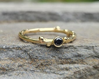 May include: A gold ring featuring a textured band that resembles a small twig or branch. The ring is set with a single, round, faceted black gemstone held in a bezel setting. The metal has a polished finish and a natural, organic appearance.