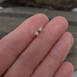 May include: A pair of small, square-shaped diamond stud earrings. The earrings are set in a gold metal and are displayed on a light-colored background. The diamonds appear to be clear and sparkling.