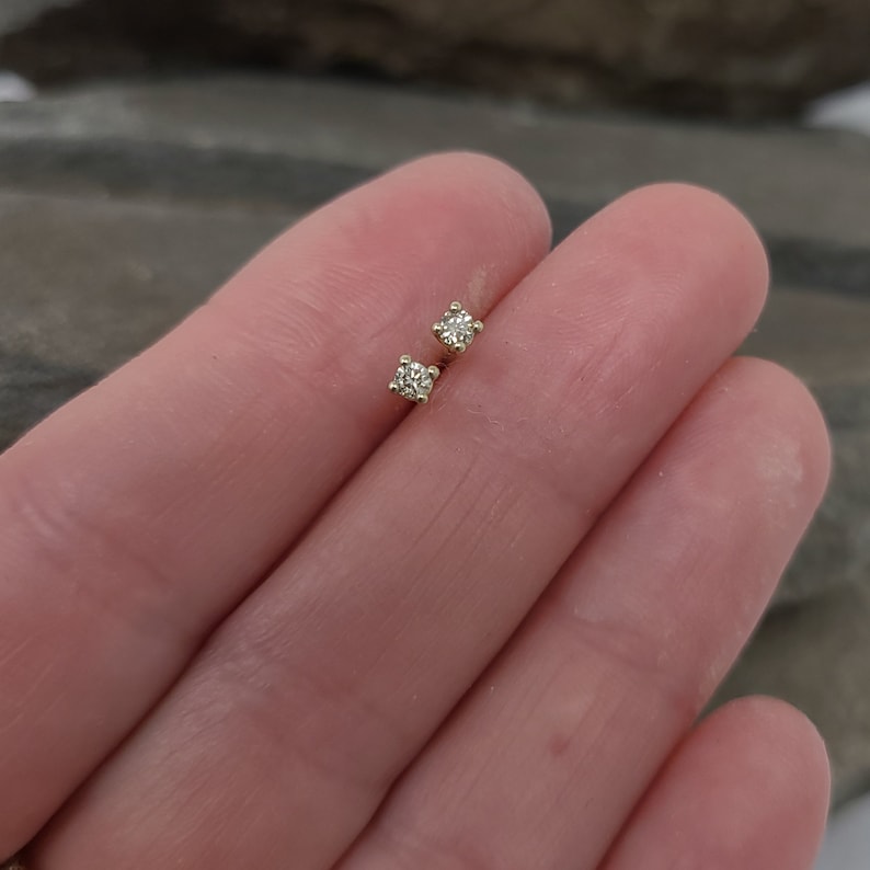 May include: A pair of small, square-shaped diamond stud earrings. The earrings are set in a gold metal and are displayed on a light-colored background. The diamonds appear to be clear and sparkling.