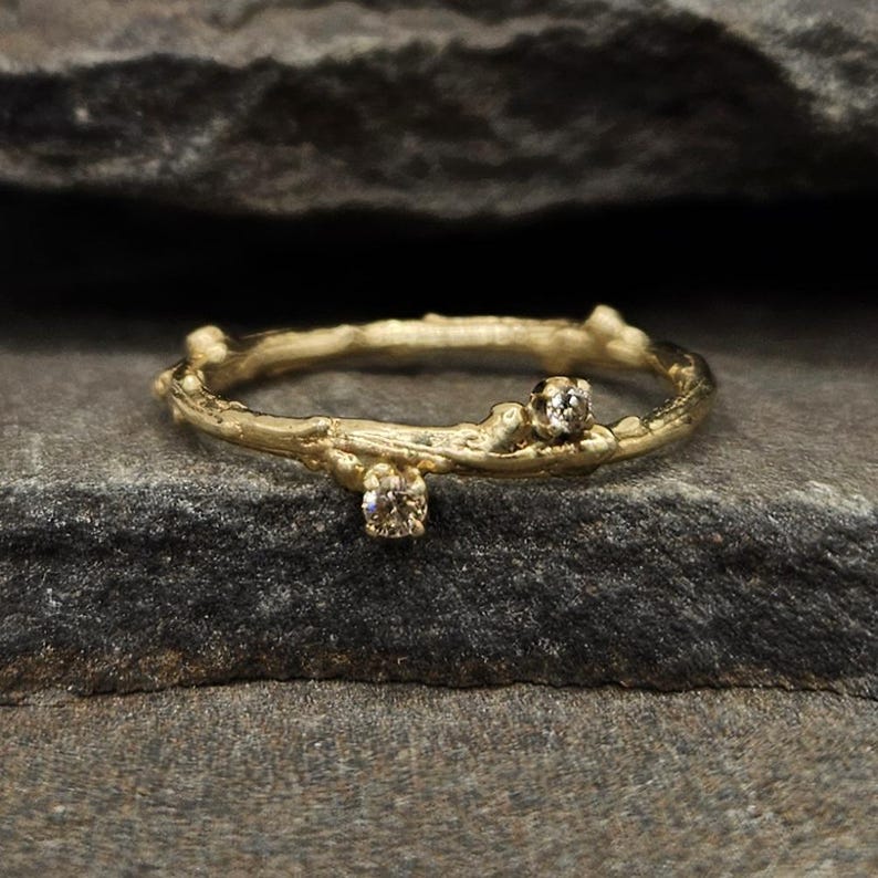 May include: A gold ring with a textured band resembling a small tree branch or twig. The band features two small, round, clear faceted stones set into the metal, positioned at different points along the circumference of the circular ring.