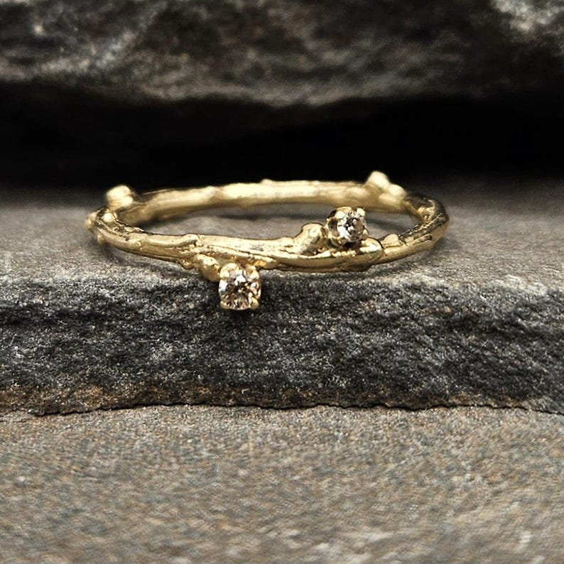 May include: A gold ring with a textured band resembling a small tree branch. The band features two small, round, clear gemstones set into the metal. The surface of the ring has an organic, uneven appearance with bumps and knots along the circumference.
