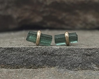May include: A pair of stud earrings featuring raw, elongated green tourmaline crystals. Each crystal is horizontally oriented and secured by a central gold-toned metal band that wraps around the middle of the stone, creating a simple, linear design.