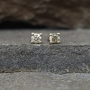 May include: A pair of small, square-set diamond stud earrings. The diamonds are clear and sparkling, set in a gold-colored metal. The earrings are displayed on a dark gray, textured surface.