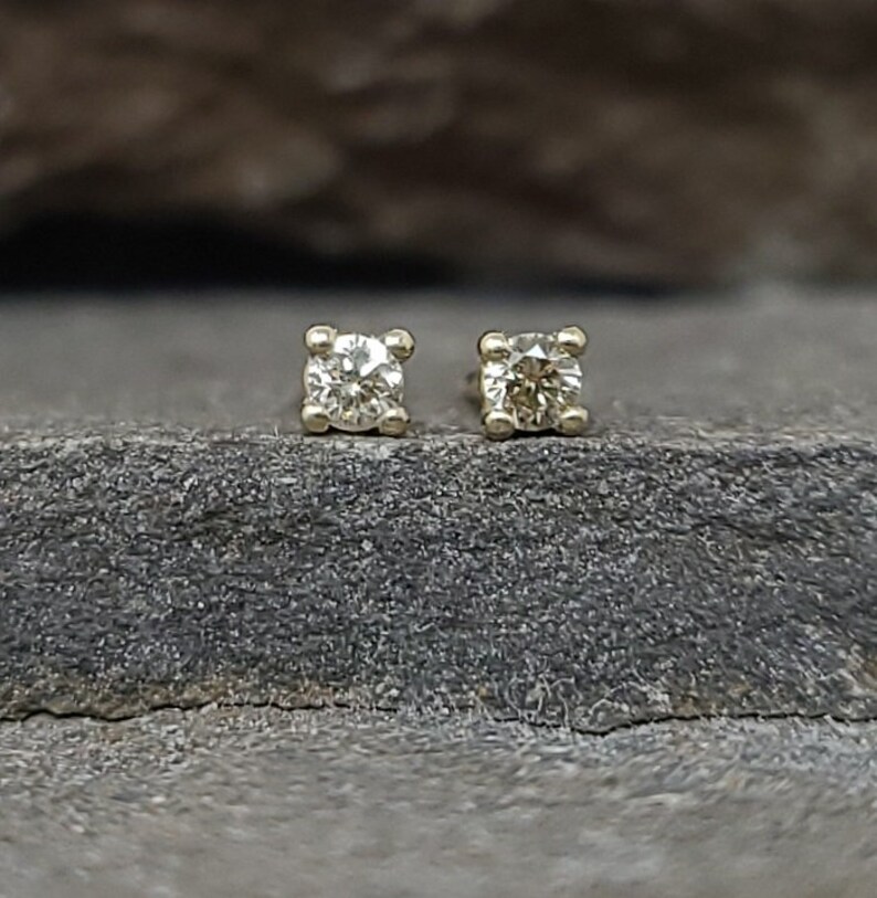May include: A pair of small, square-set diamond stud earrings. The diamonds are clear and sparkling, set in a gold-colored metal. The earrings are displayed on a dark gray, textured surface.