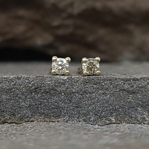 May include: A pair of small, square-shaped diamond stud earrings. Each earring features a single, round-cut diamond set in a gold or gold-toned metal. The earrings are displayed on a dark, textured surface.